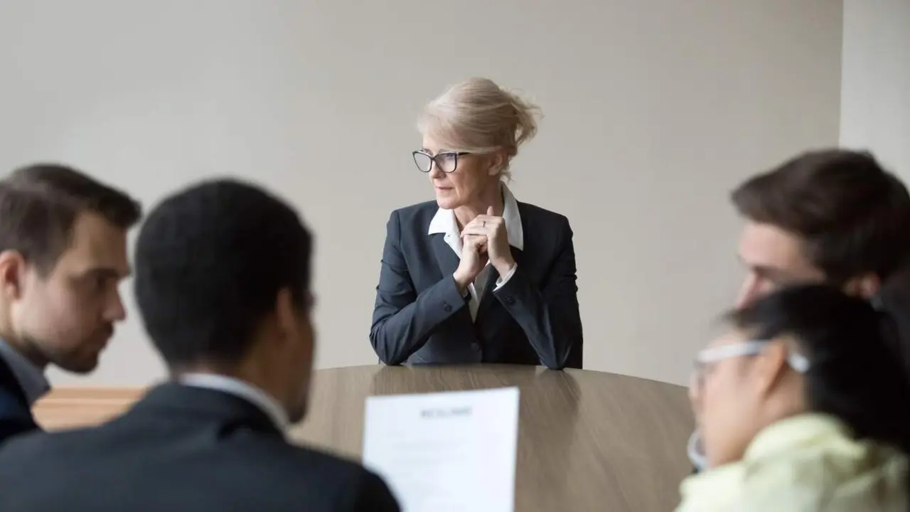 Mujer de mediana edad estresada sentada en el escritorio de la oficina durante una reuni&oacute;n. Equipo de trabajadores j&oacute;venes multirraciales sentados de espaldas a la c&aacute;mara. Concepto de entrevista de trabajo, contrataci&oacute;n, reclutamiento y discriminaci&oacute;n por edad. - Foto publicada por la Fundaci&oacute;n M&aacute;s S&eacute;nior